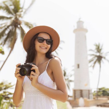 Güneş şapkalı ve güneş gözlüklü bir kadın, Sri Lanka 'daki Galle Fort deniz fenerinin önünde kamerasını tutuyor. Palmiye ağaçlarının parlak, tropik manzarası macera ve seyahat görüntülerini çağrıştırıyor..