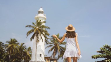 Geniş kenarlı şapkalı ve beyaz elbiseli bir kadın Sri Lanka 'daki Galle Fort deniz fenerine bakıyor. Arka planda palmiye ağaçları ile, bu güneşli tropikal sahne keşif ruhunu yakalar ve