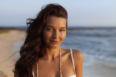 Beyaz elbiseli bir kadın günbatımında tropik bir plajda dikilirken gülümsüyor, arkasında nazik okyanus dalgaları olan bir kadın. Seyahat, yaz tatili ve sahil yaşam tarzı kavramları için ideal..