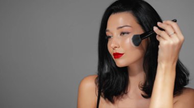 Kırmızı dudaklı, makyaj fırçası kullanan, ışıldayan tenine ve kusursuz güzelliğine odaklanmış şık bir kadının yakın çekimi. Makyaj dersleri, moda promosyonları ve cilt reklamları için mükemmel.