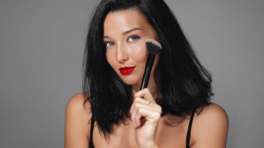 Cesur kırmızı dudakları ve kusursuz derisi olan, makyaj fırçası tutan, güzelliği ve zarafeti ön plana çıkaran kendine güvenen bir kadının yakın portresi. Kozmetik promosyonlar, cilt bakımı reklamları ve güzellik dersleri için ideal.