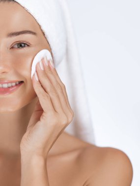 Yüzünde parlayan derisi olan gülümseyen genç bir kadın yüzünü temizlemek için pamuk ped kullanıyor. Havluya sarılmış halde, cilt bakımını temiz ve sakin bir ortamda yapmaktan hoşlanıyor. Güzellik, cilt bakımı ve sağlık için idealdir.