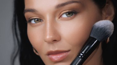 Güzel bir kadının bir fırçayla makyaj yaptığı, pürüzsüz cildini ve büyüleyici gözlerini vurguladığı yakın plan bir fotoğraf. Güzellik, cilt bakımı ya da kozmetik ürünler için mükemmel..