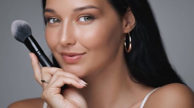 Güzel bir genç kadın elinde makyaj fırçası tutarken gülümsüyor, kusursuz cildini ve parlak tenini gösteriyor. Güzellik, cilt bakımı ve kozmetik ürünler için mükemmel..
