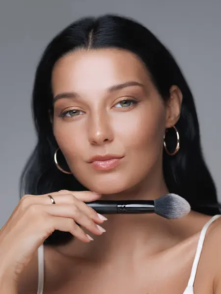 stock image A confident young woman with glowing skin holds a makeup brush under her chin, emphasizing her flawless beauty. Ideal for beauty, skincare, and cosmetic promotions or lifestyle content.