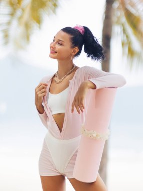 Gülümseyen bir kadın tropik plaj atmosferinin tadını çıkarırken pembe yoga minderine yaslanır. Spor kıyafetleri içinde, sakinlik ve açık hava sağlık seansı için hazır olma havası yayıyor. Tam oldu.