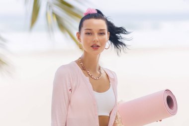 Bir kadın tropik bir plajda pembe bir yoga minderi tutuyor, açık hava fitness seansına hazırlanıyor. Rahatlamış ama kendine güvenen ifadesi farkındalığın ve sağlıklı yaşamın özünü yakalıyor. İdeal