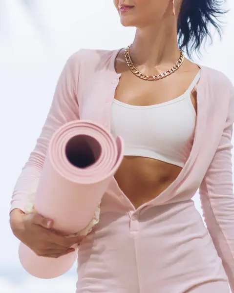 stock image A stylish woman dressed in a pink outfit and gold necklace, holding a pink yoga mat, ready for a workout session. Perfect for fitness, wellness, and active lifestyle promotions.