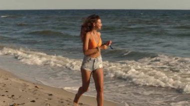 Güzel bronzlaşmış bir kız plaj boyunca turuncu bir bikini bluzu ve kot şortuyla koşuyor. Bu canlı sahne yaz eğlencesinin ve kaygısız plaj maceralarının özünü yakalıyor..