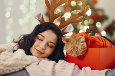 Ren geyiği boynuzları takan neşeli kadın Noel kutusundaki bir kedinin yanında dinleniyor. Konforlu tatil sahnesi mevsimlik promosyonlar, şenlikli reklamlar ve tatil temalı içerik için mükemmel.. 