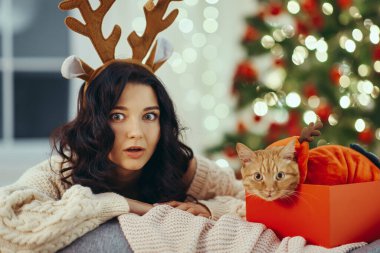 Noel kutusunda kediye geyik boynuzu takan sürpriz bir kadın. Konforlu tatil sahnesi mevsimlik promosyonlar, Noel reklamları ve neşeli tatil temalı içerik için mükemmel.