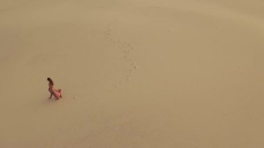 Doğada yolculuk videosu. Güzel dalgalı kırmızı elbiseli genç bir kadın doğal ortamda yürüyor. 4K Hava Aracı, rüzgarlı bir günde kum tepesinde gün batımında beyaz bir kadının görüntüsü.