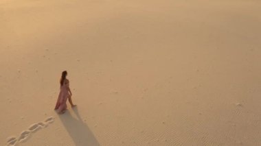 Çöldeki kum tepelerinde yürüyen pembe elbiseli muhteşem bir kadının yanında uçan bir dronun hava görüntüsü. Gezgin moda kızı sineması görüntüleri. 4K Sahne Doğası
