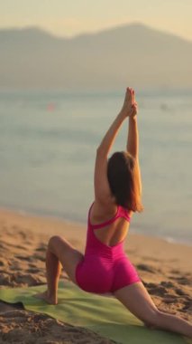 Bir kadın güneş doğarken sahile bakan bir plajda hareketli yoga pozu uyguluyor. Barışçıl, yavaş çekim sahnesi, güç ve farkındalığı vurguluyor, fitness ve sağlık promosyonları için ideal. Dikey