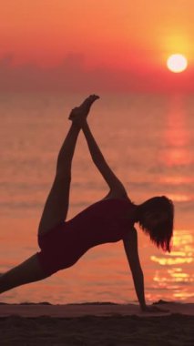 Dikey videoda bir kadın gün doğumunda plajda zarif bir yoga pozu verir ve parlak ufukta güzel bir siluet oluşturur. Bu sakin sahne esnekliği ve farkındalığı vurguluyor.