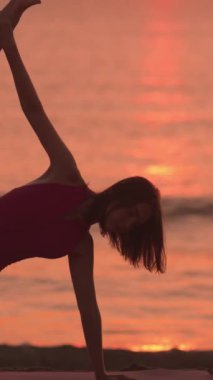 Bir kadın, gün doğumunda plajda, parıldayan ufukta siluet şeklinde esneklik odaklı yoga pozu veriyor. Huzurlu yavaş çekim sahnesi sağlığın, farkındalığın ve gücün altını çiziyor.