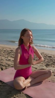 Dikey video. Pembe spor kıyafetli bir kadın, kumlu bir sahilde, parlak sabah güneşinin altında yoga pozuyla meditasyon yapıyor. Arka planda sakin bir okyanus ve dağlarla, huzurlu manzara