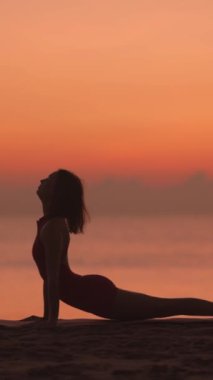 Bir kadın yoga pozları verirken siluetlenir, plajda güneş doğarken yukarıdan aşağıya doğru bir köpeğe dönüşür. Bu barışçıl sahne farkındalığı ve esnekliği vurguluyor, sağlık için mükemmel.