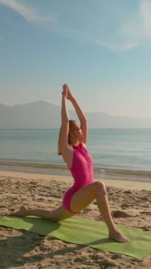 Bir kadın sabah, sakin bir plajda yoga pozu verir, okyanusa bakar ve yumuşak güneş ışığında güneşlenir. Fitness, farkındalık ve kıyı rahatlama temalarını desteklemek için idealdir. Dikey