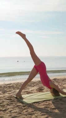 Bir kadın, arka planda sakin bir okyanusla kumlu bir plajda yükselen bir ayakla aşağı doğru köpek yogası yapıyor. Farkındalık, esneklik, sağIık ve açık hava fitness temaları için ideal