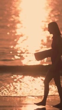 Bir kadın silueti gün doğumunda plajda yoga minderiyle yürürken görülüyor. Ağır çekim sahnesi, rahatlık ve rahatlama içeriği için ideal sabahın huzurlu güzelliğini yakalar. Dikey