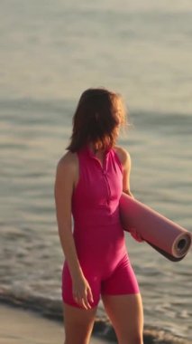 Pembe kıyafetli genç bir kadın elinde yoga minderiyle gün doğumunda kıyıda yürüyor. Bu görüntü sağlık, plaj yogası ve rahatlama kavramları için idealdir. Huzurlu bir anı yakalamak için.