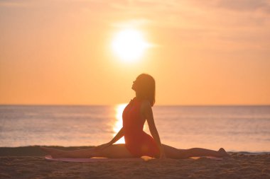 Yoga yapmış bir kadının silueti güneşin doğuşuyla plajda poz veriyor ve güneş vücudunu aydınlatıyor. Bu imaj, refahı, esnekliği, farkındalığı ve barışı teşvik etmek için idealdir.