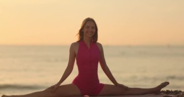 Pembe yoga kıyafeti giymiş formda bir kadın gün batımında huzurlu bir sahilde poz veriyor. Huzurlu okyanus manzarası bu sahneyi esneklik, sağIık ve zindelik için ideal hale getiriyor.. 