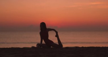 Gün batımında, arka planda okyanus olan bir kadının yoga pozunda derin bir esneme hareketi yaptığı huzurlu bir siluet. Farkındalığı, rahatlığı, yogayı ve açık havayı teşvik etmek için ideal.
