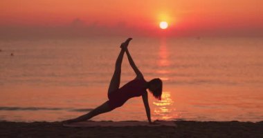 Bir kadın, gün doğumunda sahilde yoga pozu veriyor, okyanus ufkuna doğru siluet şeklinde. Zindelik, zindelik ve farkındalık için idealdir. Huzurlu ve doğal güzellikler sergilenir..