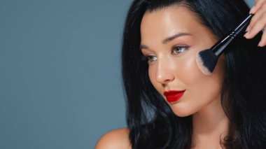 Cesur kırmızı dudakları olan, fırça kullanarak makyaj yapan, kusursuz cildini ve zarafetini sergileyen göz kamaştırıcı bir kadının yakın çekimi. Güzellik, cilt bakımı ve kozmetik promosyonları için idealdir.