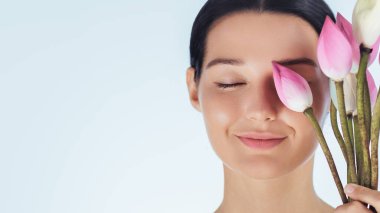 Gözleri kapalı, elinde pembe nilüfer çiçekleri tutan huzurlu genç bir kadının yakın çekimi. Yumuşak ışıklandırma ve minimalist arka plan sükunet, doğal güzellik ve refah kavramlarını iletir..