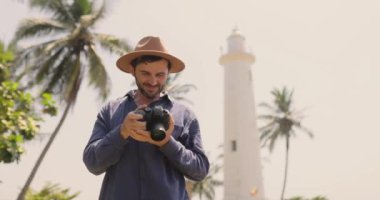 Şapkalı bir adam, Sri Lanka 'daki Galle Fort deniz fenerinin önündeki fotoğraflara bakıyor. Tropikal palmiye ağaçları ve güneşli bir gökyüzü ile çevrelenmiş bu resim huzurlu bir yolculuğu yakalar ve
