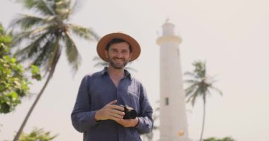 Sri Lanka 'daki Galle Fort deniz fenerinin yakınlarında elinde kamera olan, şapka dalgaları takan bir adam. Palmiye ağaçları ve parlak gökyüzü ile bu tropikal manzara seyahat, turizm ve macera için mükemmeldir.