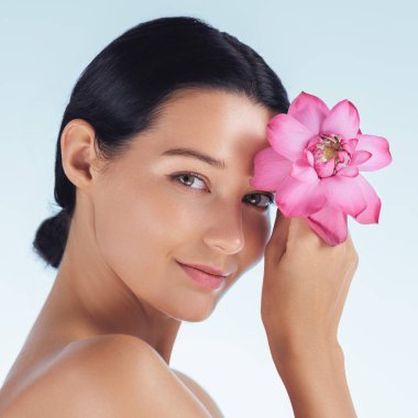 Açık tenli güzel bir kadın açık mavi arka planda pembe bir nilüfer çiçeği tutar, doğal güzellik ve cilt bakımını sembolize eder. Sağlık için ideal, spa promosyonları ya da güzellik reklamları için..