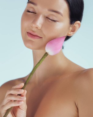 Huzurlu çıplak bir kadın nazikçe yüzüne yakın pembe bir nilüfer çiçeği tutar, saflık ve sakinliği somutlaştırır. Bu resim, kusursuz cildi ve sakin duruşuyla, cilt bakımını ve sağlığını arttırmak için mükemmel.