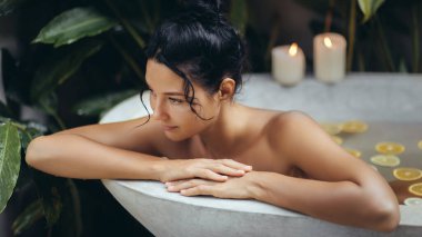 Serene young woman of Asian descent relaxes in a luxurious bathtub filled with citrus slices, surrounded by tropical plants and candlelight. Ideal for banners promoting wellness, self-care, and spa clipart