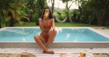 Turuncu bikinili güzel bir kadın, serin bir havuz kenarında otururken ferahlatıcı bir meyve suyunun tadını çıkarıyor. Tropikal tatilleri, yaz yaşam tarzını ve sağlık kavramlarını desteklemek için ideal.