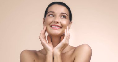 Neşeli bir gülümseme ile ışıl ışıl genç bir kadın, yakın plan bir portrede nazikçe yüzüne dokunuyor. Işıl ışıl cildi ve doğal güzelliği yumuşak bir arka plana vurgulanarak bu görüntüyü mükemmel hale getiriyor.