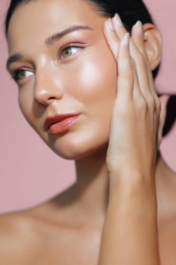 Işıl ışıl cildi ve doğal makyajı olan Latin bir kadının güzel bir yakın çekimi. Elini yumuşak pembe arka planda nazikçe yanağına yaslıyor. Cilt bakımı promosyonları, güzellik makaleleri ve...