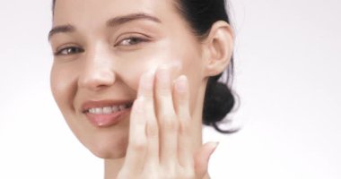 Parlak tenli sakin genç bir kadın gözleri kapalı gülümserken yüzüne krem sürüyor. Güzellik, cilt bakımı ya da hidrasyon, kendine bakım ve sağlıklı yaşam için ideal.