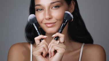 Bir kadın elinde iki makyaj fırçasıyla poz verir, kusursuz cildini ve doğal parıltısını vurgular. Güzellik, kozmetik ve cilt bakım ve makyaj dersleri üzerine odaklanan terfiler için ideal.
