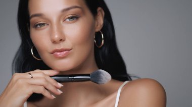 Güzel bir genç kadın kusursuz cildini ve zarif görünüşünü vurgularken makyaj fırçası sergiliyor. Bu görüntü güzellik, kozmetik ve cilt bakımı için mükemmeldir..
