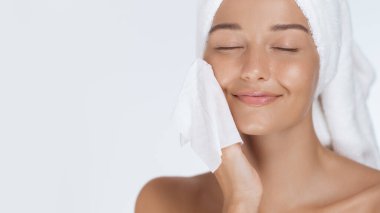 Saçına havlu sarılmış dingin bir kadın yumuşak bir havlu, gözleri kapalı ve rahat bir gülümsemeyle yüzünü nazikçe temizliyor. Güzellik, cilt bakımı ve kendini geliştirmek için mükemmel.