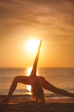 Bir kadın silueti, gün batımında kumlu bir plajda, esneklik, denge ve sakinliği ön plana çıkaran uzun bir kenar açı yoga pozu verirken yakalanır. Sağlık, yoga ve zindelik için ideal.