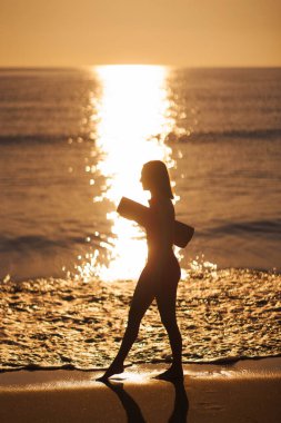Gün doğumunda bir kadın silueti, elinde yoga minderiyle kumsalda yürürken yakalanır. Bu huzurlu görüntü, sağlık, farkındalık ve sakin bir sabah rutinini çağrıştırıyor. Yoga ve zindelik için mükemmel.