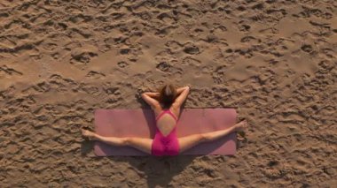 Gün batımında kumlu bir plajda yoga yapan bir kadının havadan görünüşü. Esneklik, farkındalık, açık hava fitness ve doğada barışçıl sağlık rutinleri temaları için mükemmel..