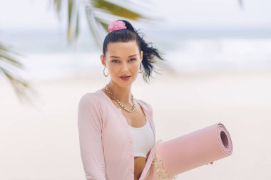 Kendine güvenen bir kadın tropikal bir plajda pembe bir yoga minderi tutuyor. Sağlık seansına hazır. Spor kıyafetleri giyer, sağlık ve rahatlığı somutlaştırır ve bunu terfi için mükemmel kılar.