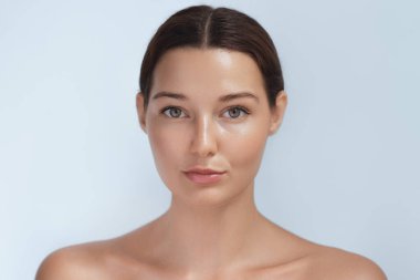 Doğrudan kameraya bakan parlak, doğal ciltli genç bir kadının portresi. Yumuşak ışıklandırma ve minimalist arka plan kusursuz cildini vurgular, güzellik ve cilt bakımı için mükemmeldir.