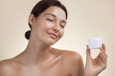 Cildi parlayan gülümseyen genç bir kadın bir kavanoz cilt kremi tutuyor, gözleri kapalı, mutluluk saçıyor. Güzellik ürünlerini, cilt rutinlerini, sağlık ve kendine bakımı teşvik etmek için mükemmel.